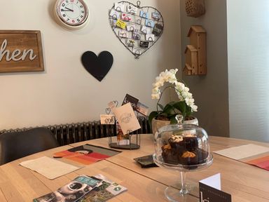 A dining table with cupcakes under a glass dome and a white orchid plant in a pot on it with wall clock and heart shaped decor on the wall at Hillcroft in St Austell