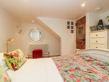 A bedroom with a bed with floral pillows and quilt a red ottoman a round mirror a wooden door and a white dresser at Hillcroft in St Austell