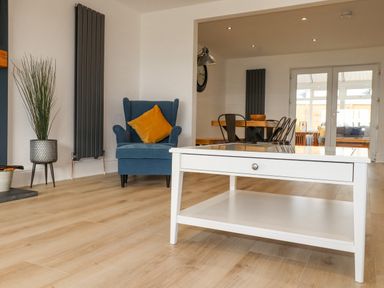 A living room with a blue armchair with a yellow cushion a white coffee table and a dining area in the background at Tarth Y Mor in Criccieth