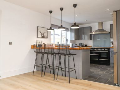 A kitchen with a wooden breakfast bar and three black wire stools under hanging lights at Tarth Y Mor in Criccieth