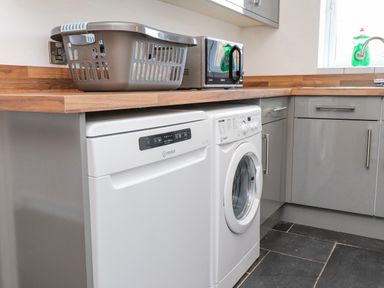 A kitchen with a washing machine and dishwasher under a wooden countertop with a laundry basket and microwave on top at Tarth Y Mor in Criccieth