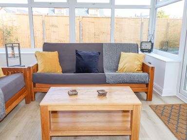 A seating area with wooden sofas, cushions, a wooden coffee table, and a clock on the windowsill at Tarth Y Mor in Criccieth