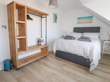 A bedroom with a double bed with white bedding and gray headboard beside a wooden open wardrobe and white bedside tables at Tarth Y Mor in Criccieth