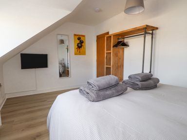 A bedroom with a bed and folded towels a wall-mounted TV a full-length mirror and an open wooden wardrobe at Tarth Y Mor in Criccieth