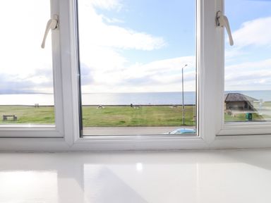 A view of a grassy area with benches and a shelter near the sea seen through a white window frame at Tarth Y Mor in Criccieth
