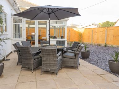 An outdoor patio with a round glass table surrounded by eight gray wicker chairs and a large umbrella at Tarth Y Mor in Criccieth