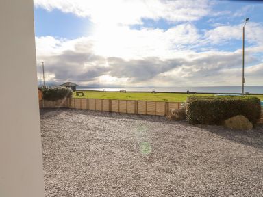 A gravel area with a wooden fence hedge and rock overlooking a grassy area benches and a sea view at Tarth Y Mor in Criccieth