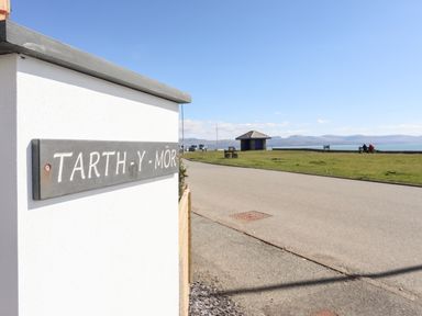 A road with a small pavilion and people sitting on benches near a grassy area by the sea at Tarth Y Mor in Criccieth