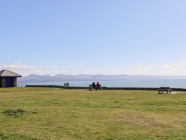 A grassy open area with a bench and picnic table overlooking the sea and distant mountains at Tarth Y Mor in Criccieth