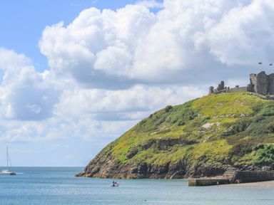 A coastal scene with a grassy hill and castle ruins overlooking the sea with boats and people on the shore at Tarth Y Mor in Criccieth