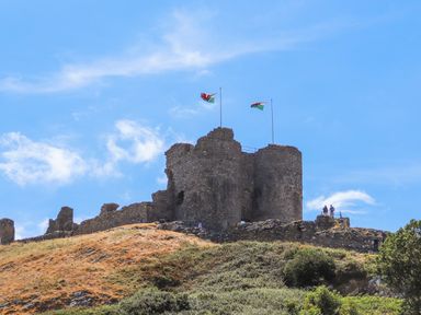 A stone castle ruin on a hill with two flags flying and people standing near the edge at Tarth Y Mor in Criccieth