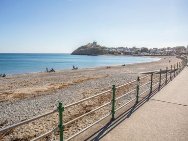 A pebble beach with a railing and a castle on a hill in the background at Tarth Y Mor in Criccieth