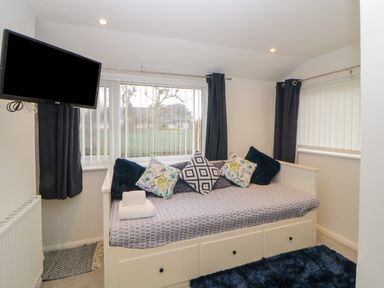 A small bedroom with a white daybed with patterned cushions a wall-mounted TV and windows with vertical blinds at Cornerside in Sidmouth