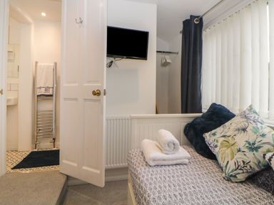 A bedroom with a bed with pillows and folded towels next to a window with blinds and a door leading to a bathroom with a towel on a heated rail at Cornerside Sidmouth