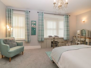 A bedroom with a bed a teal armchair with a cushion curtains and a dressing table at Cornerside in Sidmouth