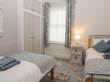 A bedroom with two single beds a window with vertical blinds and blue floral curtains a bedside table with a lamp and built in wardrobes at Cornerside in Sidmouth