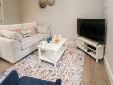 A living room with a white sofa pink and patterned cushions a white coffee table with mugs and pastries a flat screen tv on a white tv stand and a patterned rug at 3 Compass Point in Weymouth