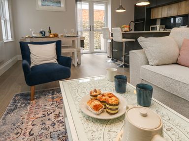 A living area with a beige sofa and blue armchair around a white coffee table with muffins and cups in a kitchen dining space at 3 Compass Point in Weymouth