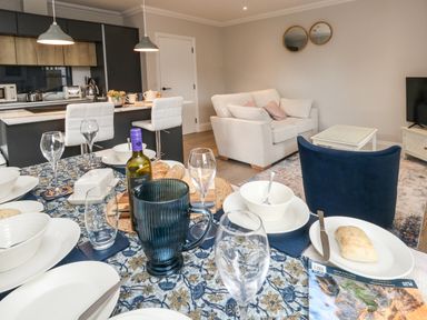 A dining table set with plates glasses and cutlery next to a kitchen with bar stools and a living room with a sofa and armchair at 3 Compass Point in Weymouth
