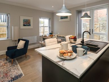 A kitchen island with cups and pastries and a bowl of fruit next to a dining table set with dishes and glasses and a blue armchair at 3 Compass Point in Weymouth