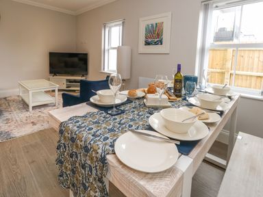 A dining table set with bowls, plates, glasses, bread, and a bottle next to a living area with a television and blue chairs at 3 Compass Point in Weymouth