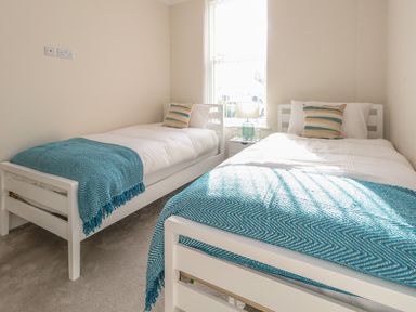 A bedroom with two single beds with blue blankets and striped pillows with a window and a bedside table with a lamp at 3 Compass Point in Weymouth