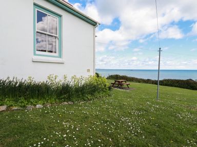 Side of a white house with a window and garden next to a lawn with a picnic table and a sea view at Beachcliff in Gorran Haven