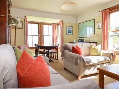 A living room with two sofas with colorful cushions a wooden dining table near windows and a sideboard with a lamp and radio at Beachcliff in Gorran Haven