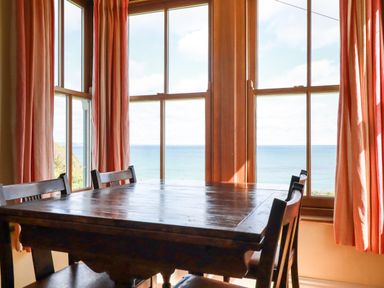 A dining area with a wooden table and chairs by large windows overlooking the sea at Beachcliff in Gorran Haven