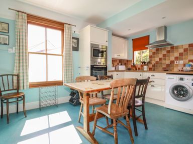 A kitchen with a wooden table and four chairs a washing machine and an oven next to a window with curtains at Beachcliff in Gorran Haven