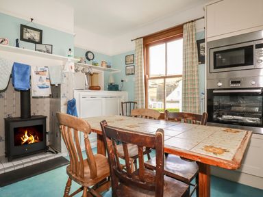 A kitchen with a tiled dining table surrounded by wooden chairs a wood burning stove and a window with checkered curtains at Beachcliff in Gorran Haven