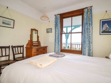 A bedroom with a bed and folded towels a wooden dresser with mirror two chairs and a window with blue curtains at Beachcliff in Gorran Haven
