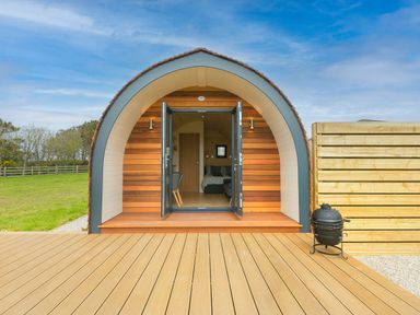 A wooden pod structure with open double doors leading to an interior with a bed and chairs on a deck at Sun Rise in St Buryan