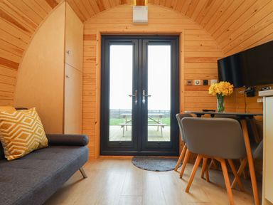 A small wooden room with a grey sofa and yellow cushion beside a cabinet and glass doors with a table and chairs on the right with a vase of flowers and a TV mounted on the wall at Sun Rise in St Buryan