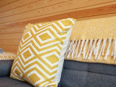 A yellow and white patterned cushion and a yellow and white fringed blanket on a grey sofa with a wooden wall behind at Sun Rise in St Buryan