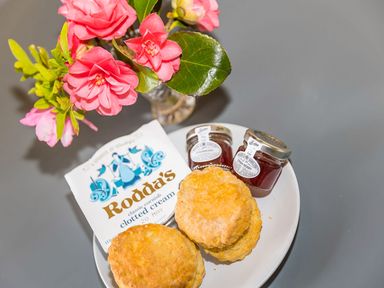 A plate with two scones small jars of strawberry jam and a packet of clotted cream with a vase of pink flowers