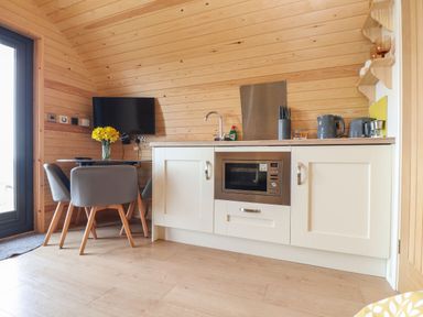 A small kitchen area with white cabinets a microwave a sink a kettle and a table with chairs and flowers at Sun Rise in St Buryan