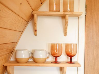 Two ceramic mugs and two orange glass goblets on a wooden shelf with wood paneling and a door in the background at Sun Rise in St Buryan