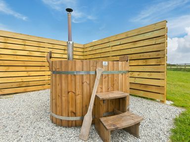 A wooden hot tub with a metal chimney on a gravel surface next to a wooden privacy fence and grassy field at Sun Rise in St Buryan