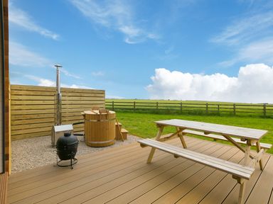 An outdoor wooden deck with a picnic table a wooden hot tub a black grill and a wooden fence with a grassy field beyond at Sun Rise in St Buryan