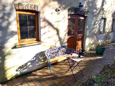A patio with a wooden bench and a small table holding two glasses and a bottle in front of a house with wooden windows and doors at Nook Bethesda near Narberth