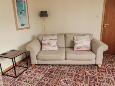 A beige sofa with patterned cushions next to a small side table and standing lamp in a room at Causeway Cottage in Holy Island