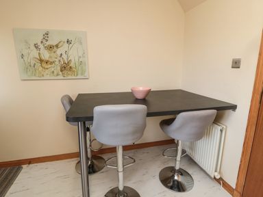 A dining area with a black table, three gray bar stools, a pink bowl on the table, and a rabbit painting on the wall at Causeway Cottage in Holy Island
