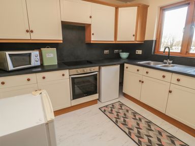 A kitchen with white cabinets black countertops a microwave oven a stove a small refrigerator a window and a patterned rug on the floor at Causeway Cottage in Holy Island