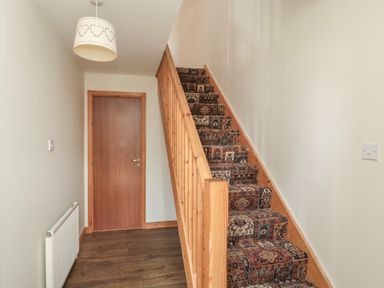 A hallway with a wooden staircase covered in patterned carpet next to a closed wooden door at Causeway Cottage in Holy Island