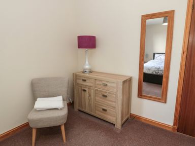 A corner with a wooden dresser and a lamp next to a gray chair with folded towels and a wall mirror showing a bed at Causeway Cottage in Holy Island