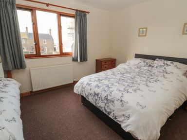 A bedroom with a double bed and another single bed with butterfly patterned bedding a wooden dresser and a window with grey curtains at Causeway Cottage in Holy Island
