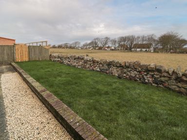 A garden with grass bordered by a stone wall and a wooden fence with houses and trees in the distance at Causeway Cottage in Holy Island