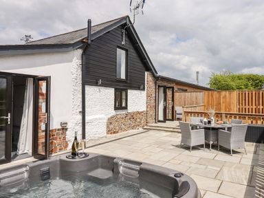 An outdoor patio with a hot tub a table with chairs and a house with open doors at Strawberry Bank - Kingfisher in Pyworthy near Bridgerule