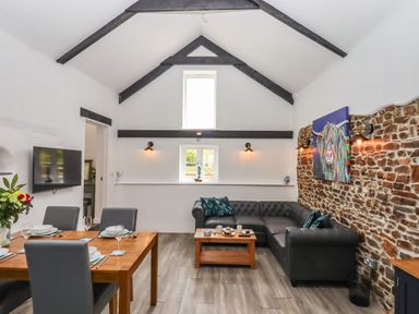 A living room with a dining table set for four and a black sectional sofa against a stone wall at Strawberry Bank - Kingfisher in Pyworthy near Bridgerule